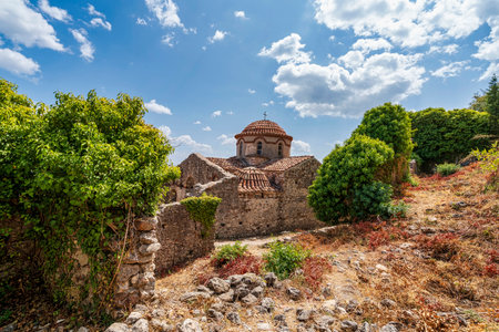 The ancient Church of St. Nicholas in Mystras, Greece, surrounded by lush greenery, stone ruins, and...