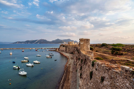 A stunning view of Methoni Fortress' coastal walls overlooking the calm harbor, with moored boats and scenic mountains in the background.の写真素材