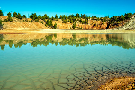 A shallow artificial lake in an abandoned quarry in Evia, Greece, reflecting red clay hills, pine trees, and a partly cloudy sky on its cracked water surface.の写真素材