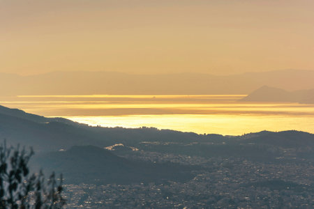View from Hymettus mountain showing Athens city with shimmering sea and distant silhouettes of islands under warm sunset light.の写真素材