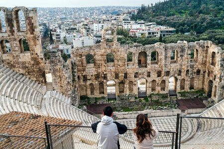 Tourists overlooking the ancient Roman amphitheater Odeon of Herodes Atticus on the Acropolis with Athens cityscape beyond.の写真素材