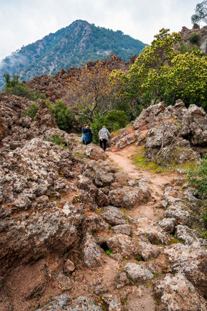 Three people hiking through rugged volcanic terrain on Kameno Vouno in Methana, surrounded by dramatic rocks and sparse vegetation.の写真素材
