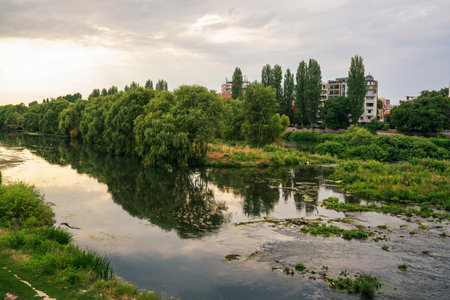 A peaceful view of the Maritsa River running through Plovdiv, Bulgaria, with lush green trees reflecting in the calm water. Modern apartment buildings line the distant bank, contrasting nature with urban life under a cloudy sky.の写真素材