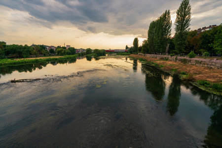 Wide-angle view of the Maritsa River in Plovdiv, Bulgaria, featuring reflections, a riverside walkway, and a distant city bridge.の写真素材
