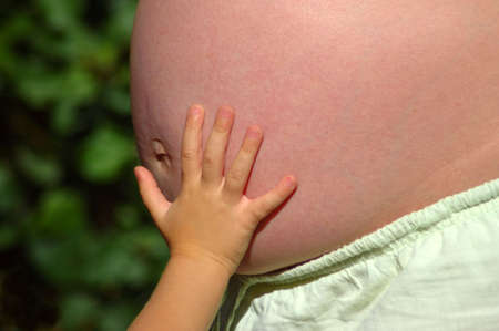 The white little hand of a caucasian toddler child touching the pregnant belly of the mother in...