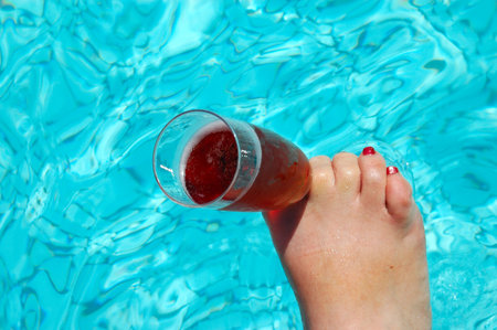 A white female foot of a caucasian lady with red nailpolish holding a champagne glass with sparkling wine in a swimming-pool の写真素材
