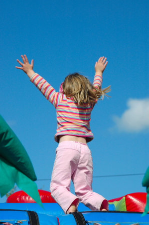 Rear view of a white caucasian girl child with long blond hair raising up her little hands in the air having great fun by jumping on a jumping castle on the playground outdoors in sunshine の写真素材