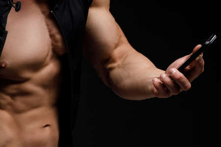 Muscular bodybuilder with phone in hands on a black background. Sports nutrition. Bodybuilding...