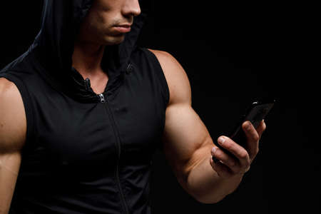 Muscular bodybuilder with phone in hands on a black background. Sports nutrition. Bodybuilding nutrition supplements, workout, healthy lifestyle concept.の写真素材
