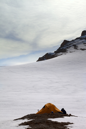 Hiker sitting near orange camping tent in evening snow mountains on overnight stay. Turkey, Central Taurus Mountains, Aladaglar (Anti-Taurus), plateau Edigel (Yedi Goller)の写真素材