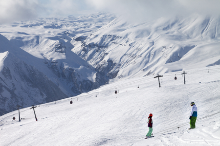 Two snowboarders descend on snowy ski slope and gondola lift at sun cloudy day. Georgia, region Gudauri. Caucasus Mountains in winter.の写真素材