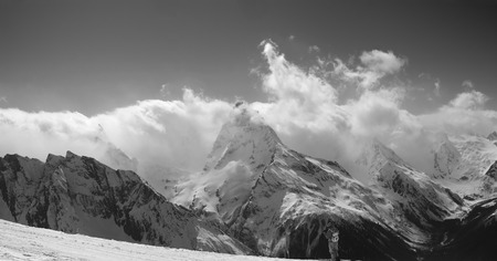 Panorama of snowy mountain peaks in sunlight clouds and ski slope with skier. Caucasus Mountains in winter, region Dombay. Black and white toned landscape.の写真素材