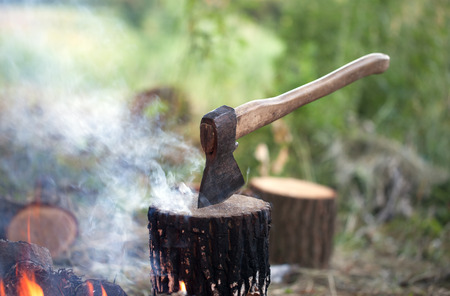 Axe in tree stump and campfire with smoke in summer forest at sunny dayの写真素材
