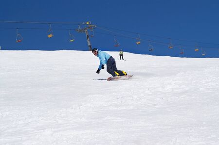 Snowboarder descends on snowy ski slope at winter mountains in sunny day. Chairlift and blue sky with snow at background. Caucasus Mountains, region Dombay.の写真素材