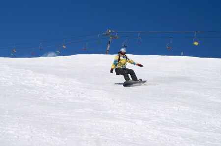 Snowboarder downhill on snowy ski slope at winter mountain in sunny day. Old chairlift and blue sky with snow at background. Caucasus Mountains, region Dombay.の写真素材