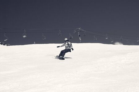 Snowboarder descends on snowy ski slope at winter mountain in sunny day. Old chairlift and sky with snow at background. Caucasus Mountains, region Dombay. Black and white retro toned image.の写真素材