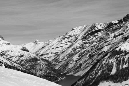 Off-piste ski slope, snowy high mountains with forest, big lake and cloudy sky at winter. Italian Alps. Livigno, region of Lombardy, Italy, Europe. Black and white toned landscape.の写真素材
