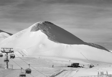 Snowy ski track prepared with snow grooming machine, chair-lift, snow cannons and snowcat at ski resort. Mountains in sunny winter day. Italian Alps. Livigno, region of Lombardy, Italy, Europe. Black and white toned landscape.の写真素材
