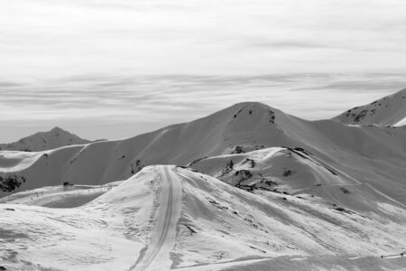 High winter mountains with ski trace, road in snowy slopes and sunlit cloudy sky at morning. Italian Alps. Livigno, region of Lombardy, Italy, Europe. Black and white toned landscape.の写真素材