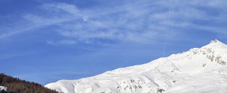 Snowy off piste slope in high winter mountains and cloudy sunlit blue sky with moon at morning. Italian Alps. Livigno, region of Lombardy, Italy, Europe. Panoramic view.の写真素材