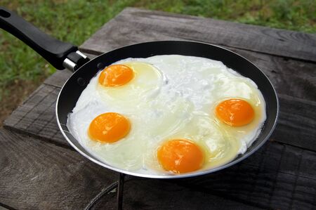 Old wooden table and frying pan with fried eggs on camping gas stove. Outdoor cooking. の写真素材