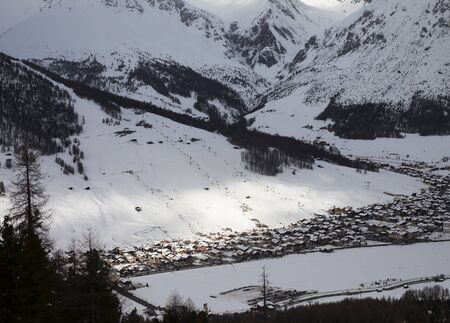 Snowy ski slope with cableways and town in mountain valley at winter evening. Italian Alps. Livigno, region of Lombardy, Italy, Europe. Top view from Mottolino ski area.の写真素材
