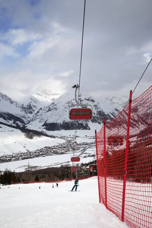 Chair-lift, snowy ski slope with snowboarders and skiers in high mountains and cloudy sunlit sky at winter. Ski area Mottolino Fun Mountain, Italian Alps. Livigno, Lombardy, Italy, Europe.の写真素材