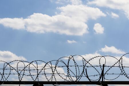 Black coiled barbed wire fence and blue sky with sunlit clouds at backgroundの写真素材