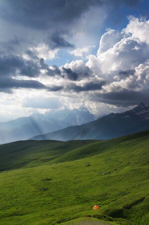 Orange tourist tent on green meadow in high mountains and sunlit cloudy sky. Caucasus Mountains. Georgia, region Svanetia in summer. の写真素材