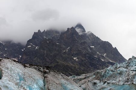 High rocky mountains in fog and glacier with deep danger crevasses covered with snow and ground at gray day. Caucasus Mountains. Georgia, region Svanetia.の写真素材
