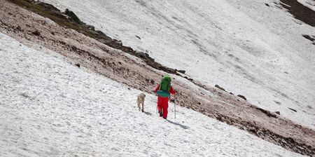 Hiker in red with dog on snowy glacier and dirty avalanche trace at high mountains. Panoramic view.の写真素材