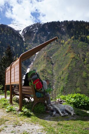 Hikers with big backpacks resting on wooden chair and dog sleeps near legs in shadow, mountains with forest and snow at background. Turkey, Kachkar Mountains in spring.の写真素材