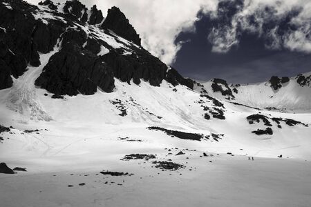 High sunlit mountains with snow cornice and avalanche track, snowy plateau and two small silhouettes of hikers at evening. Turkey, Kachkar Mountains, Pontic Mountains. Black and white toned landscape.の写真素材