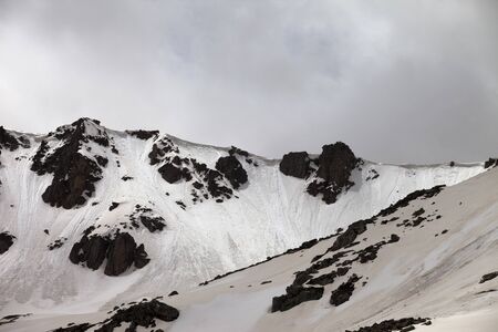 Rocks with snow cornice and traces from avalanche. High snowy mountains and cloudy sky in gray day. Turkey, Kachkar, highest part of Pontic Mountains.の写真素材