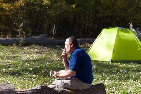 Man sitting on wooden log with mug of tea and cigarette. Hiker basks in sun, drinks tea and smokes cigarette. Sunlit light green camping tent and autumn forest at background.の写真素材