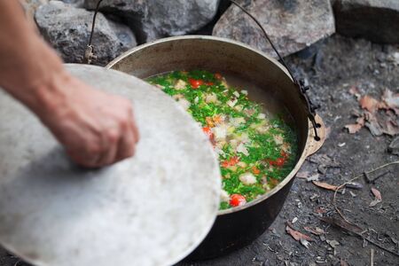 Freshly cooked hot soup in old big sooty cauldron on camp fire and man hand with pot cover. Outdoor camping cooking. Selective focus.の写真素材