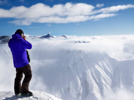 Skier in blue jacket at top of mount talking on mobile phone and snowy mountains in fog. Caucasus Mountains in sun winter day, Georgia, region Gudauri.の写真素材