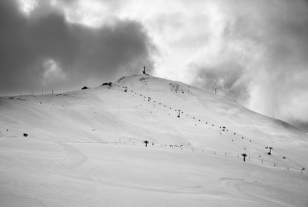 Snowy ski slope, chair-lifts in high mountains and cloudy storm sky at winter before blizzard. Ski area Mottolino, Italian Alps at bad weather. Livigno Lombardy, Italy Europe. Black and white toned.の写真素材