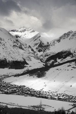 Snowy ski slope with cableways and town in mountain valley at sunny winter evening. Italian Alps. Livigno, region of Lombardy, Italy, Europe. Top view from Mottolino ski area. Black and white tonedの写真素材