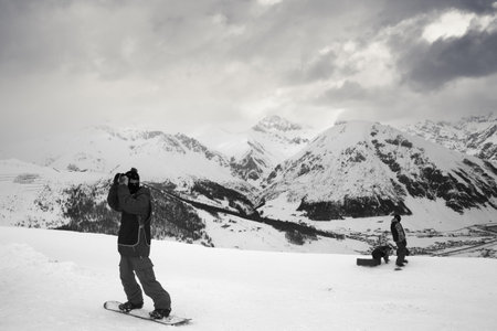 Group of snowboarders starts on off-piste descent. High snowy mountains and cloudy storm sky at winter evening. Ski area Mottolino, Italian Alps. Livigno Lombardy, Italy, Europe. Black and white tonedの写真素材