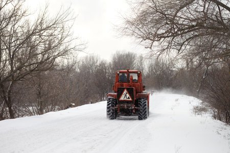 Old tractor clears the snow-covered road from snow blockages. Snowy country road through forest in bad weather after snowfall.の写真素材