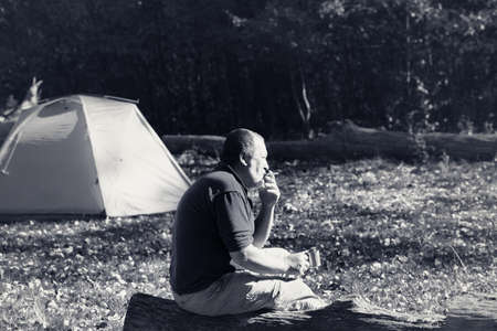 Man sitting on wooden log with mug of tea and cigarette. Hiker basks in sun, drinks tea and smokes cigarette. Sunlit camping tent and autumn forest at background. Black and white retro toned image.の写真素材