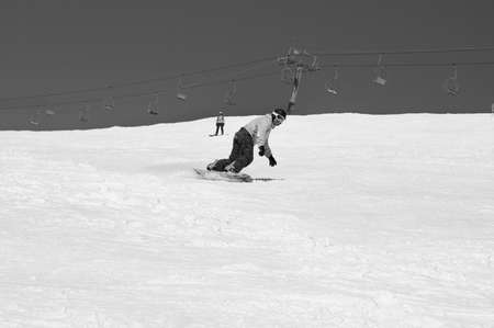 Snowboarder descends on snowy ski slope at winter mountains in sunny day. Chairlift and sky with snow at background. Caucasus Mountains, region Dombay. Black and white toned image.の写真素材