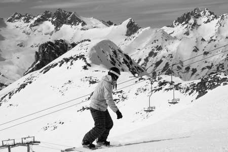 Snowboarder downhill on snowy ski slope and old chair lift in high mountains at sunny winter day. Caucasus Mountains, region Dombay. Black and white toned image.の写真素材