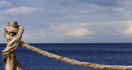 Old rope knotted on wooden post in seafront, blue sea and cloudy sky in sun summer day. Panoramic...