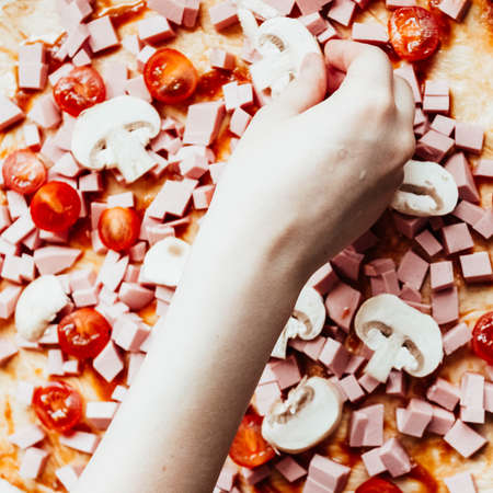 homemade pizza step by step instruction.DIY pizza step 6.Teenage girl's hand putting slice of tomato on raw pizza with tomato, sausage and mushrooms ready for baking.の写真素材