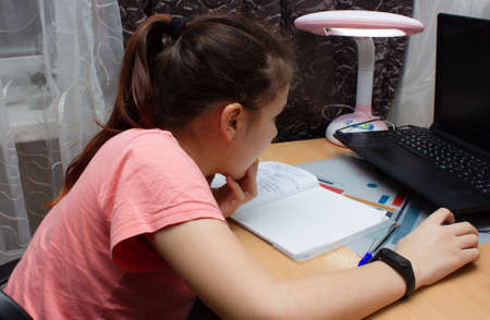 Smart Little Girl Does Homework in Her Living Room. Shes Sitting at Her Desk Writes with a Pen in...