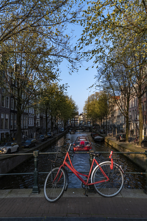 Red bicycle parking on the bridge over the calm water of a canal with a boat giving a round trip to tourists, Amsterdam, Netherlandsの写真素材
