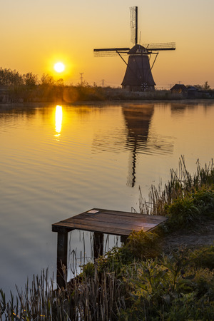 Embarquement at the edge with the calm water in the long canal during facing a windmill reflection in the burning sunrise color morningの写真素材