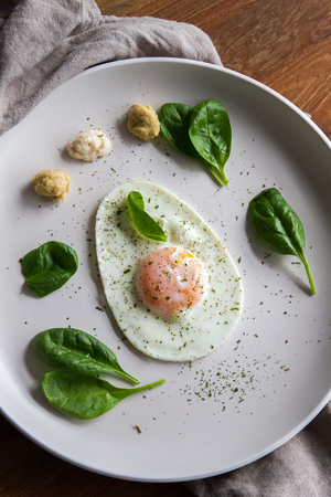 Single fried egg in egg shape over white plate garnished with spinach, green herbs, salt, papper, horseradish and hummus over wooden bord with grey napking on the sideの写真素材